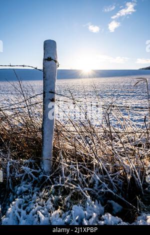 Ein alter Stacheldrahtzaun mit einem alten verfaulten Holzzaunpfosten in einem Feld, das von Morgenschnee bedeckt ist, mit einem blauen Himmel und Sonnenschein in den Cotswolds. Stockfoto