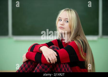 Teenager im höheren Schulalter. Ein Mädchen im Teenageralter sitzt auf dem Hintergrund einer Tafel und schaut auf die Kamera. Stockfoto