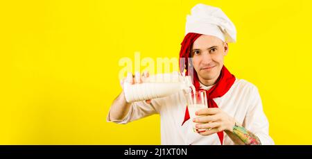 Mann mit roten Dreadlocks und Flaschenmilch. Selbstbewusste junge Köchin in weißem Hemd und roter Krawatte gießt Milch aus einer Flasche in ein Glas gegen gelben Backgro Stockfoto