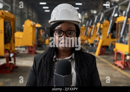 Portrait eines afrikanischen Journalisten in Arbeit Helm, der frische Nachrichten an das Mikrofon im Werk spricht Stockfoto
