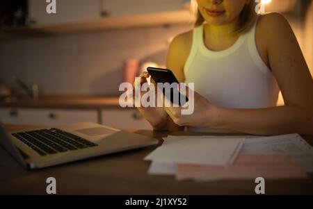 Ein fleißiges Mädchen arbeitet von zu Hause aus auf ihrem Laptop und überprüft ihr Telefon, und neben ihr befindet sich ein Stapel Papiere Stockfoto