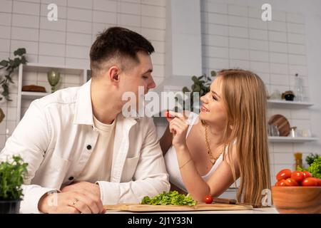 Verheiratetes Paar, das zu Hause kocht. Schöne Frau füttert Ehemann Tomate, während Gemüse Salat. Glückliche Familie, die Zeit zusammen verbringt. Hochwertige Fotos Stockfoto