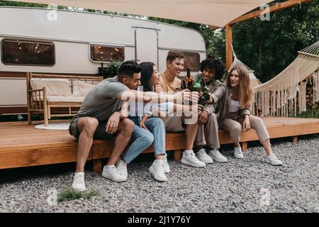 Fröhliche junge multirassische Freunde trinken Bier auf Campingausflug, toasten mit Flaschen, Spaß zusammen im Freien Stockfoto