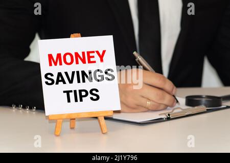 Männliche Hand, die einen Block mit dem Wort „Geld“ auf einen Block mit den Worten „Geld sparen Tipps“ setzt. Schöner Holztisch, weißer Hintergrund. Speicherplatz kopieren. Stockfoto