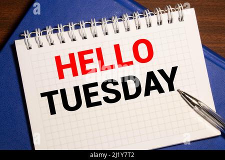 HELLO TUESDAY in rot auf einem hellen Hintergrund neben einem Computer, einem Stift, einer Lupe, einer schwarzen Brille und einer grünen Pflanze geschrieben. Motivierende Co Stockfoto