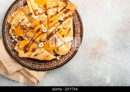 Crepes-Suzette mit Orangen und Schokolade. Dünne Crepes mit Schokoladenaufstrich, Haselnüssen und Orangenscheiben Obst auf dem Teller zum Frühstück auf altem Beton Stockfoto