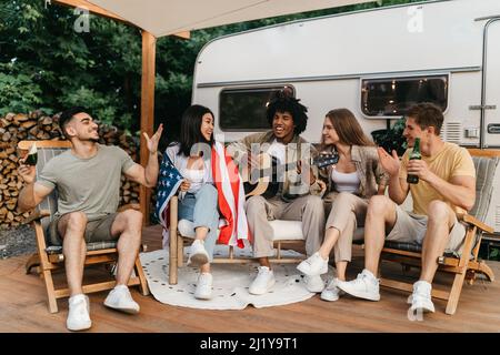 Interrassische Freunde, die Gitarre spielen und singen, amerikanische Flagge halten, Bier trinken, in den Sommerferien in der Nähe von RV sitzen Stockfoto