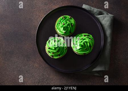 Drei Schokoladen-Cupcakes mit grüner Schlagsahne und Schokoladenstreuseln auf braunem Hintergrund. St. Patrick's Day. Speicherplatz kopieren. Blick von oben. Stockfoto