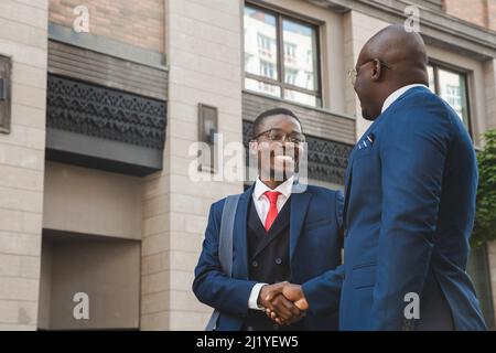 Zwei schwarze afroamerikanische Geschäftsleute in Anzügen und Gläsern schütteln sich die Hände, während sie sich im Freien treffen Stockfoto