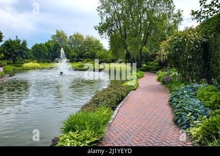 Ein Teich mit Wasser gefüllt mit einem Sprühstrahl-Brunnen in einem Park mit Fußgängerwegen aus Steinfliesen zwischen verschiedenen Pflanzen, Landschaftsgestaltung von Stockfoto