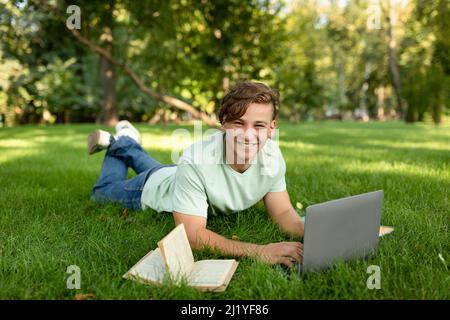 Glücklicher männlicher Student, der einen Laptop benutzt, im Freien online lernt, auf dem Rasen auf dem College-Campus oder im Park liegt Stockfoto