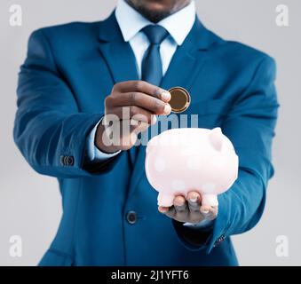 Die heutigen Ersparnisse sind das Geld der Bank von morgen. Studioaufnahme eines Geschäftsmannes, der ein Sparschwein hält und vor grauem Hintergrund eine Bitcoin in die Bank setzt Stockfoto