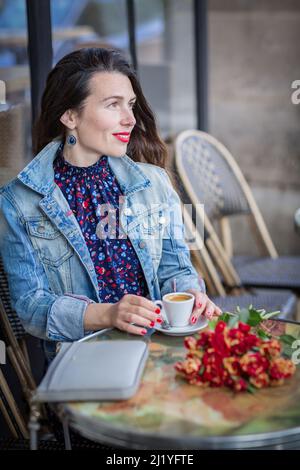 Schöne Frau mit langen brünetten Haaren in einem blauen Kleid, die allein mit Blumen im Straßencafé sitzt und Kaffee trinkt Stockfoto