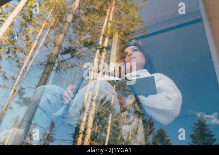 Porträt einer alleinstehenden Frau, die mit offenem Buch auf dem Balkon des Landhauses sitzt, Waldblick, frische Luft und Bestseller-Roman genießt. Hohe Fichten r Stockfoto