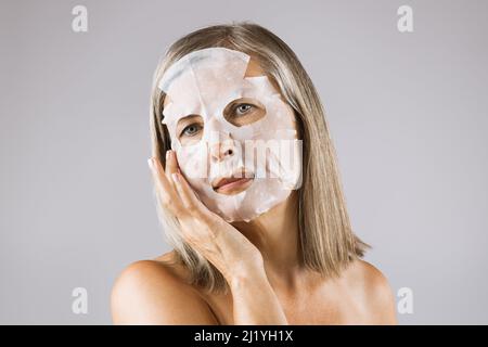 Atemberaubende kaukasische Dame mit natürlichen Falten Anwendung Papiermasken Blatt auf Gesicht im Studio. Gealterte Dame genießt Hautpflegeverfahren auf grauem Hintergrund. Stockfoto