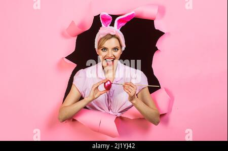Frohe Ostern. Lächelndes Mädchen in Hasenohren, das Eier malt. Kaninchenfrau mit rosa Ei und Pinsel. Stockfoto