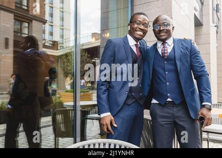 Zwei dunkelhäutige afroamerikanische Geschäftsmänner, Freunde in Anzügen, umarmen sich und freuen sich über ein Treffen auf der Straße in der Nähe eines Cafés Stockfoto