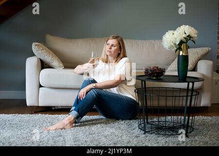 Modische Frau. Blonde kaukasische Frau mit einem Glas Wein zum Fenster schauend, während sie auf dem Teppich auf dem Boden neben dem Sofa im Wohnzimmer sitzt. Nachdenkliche Dame, die sich zu Hause ausruhte Stockfoto