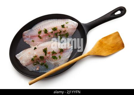 Zubereitete rohe weiße Fischfilets, die mit rosa Salz und Kräutern in einer gusseisernen Pfanne und einem Holzspatel zum Braten bestreut wurden. Isoliert auf weißem Hintergrund Stockfoto