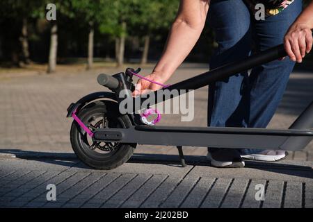 Eine unkenntliche Frau, die ihren Elektroroller an einem sonnigen Morgen in einem Stadtpark entfaltet. Konzepte von elektrischer Energie, grüner Mobilität, Umweltfreundlichkeit Stockfoto