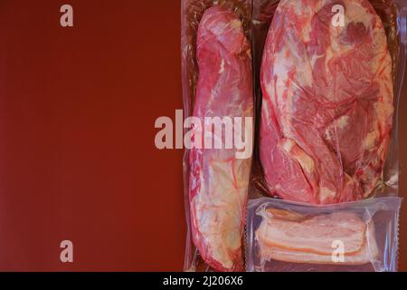 Fleisch und Schweinefett in Vakuumbeuteln auf burgunderrotem Hintergrund.Rotes Fleisch auf rotem Hintergrund. Fleischerzeugnisse. Proteinernährung. Stockfoto