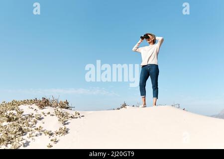 Es gibt so viel zu sehen. Ganzkörperaufnahme einer attraktiven reifen Frau, die ihr Fernglas benutzt, während sie am Strand steht. Stockfoto