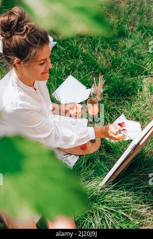 Künstler malen auf der Staffelei im Garten. Open-Air-Kunstwerkstatt. Zeichnen Sie auf der Leinwand mit Pinsel und Palette sitzen auf dem Gras während eines Picknicks in einem Park. Kreativität, Kunst und Hobby-Konzept Stockfoto