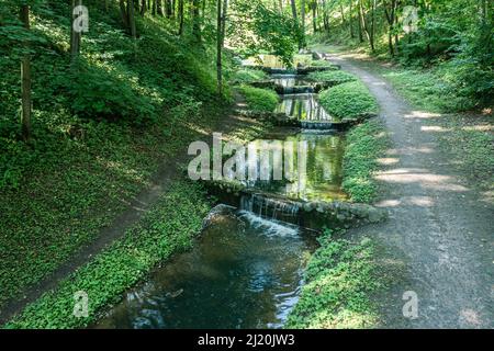 Kleine künstliche Wasserfälle im öffentlichen Park an sonnigen Sommertagen. Luftaufnahme von fliegender Drohne. Stockfoto