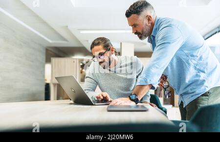 Moderne Geschäftsleute, die während der Zusammenarbeit in einem Co-Working-Büro eine wichtige Diskussion führen. Zwei fokussierte Geschäftsleute, die während der Arbeit einen Laptop benutzen Stockfoto