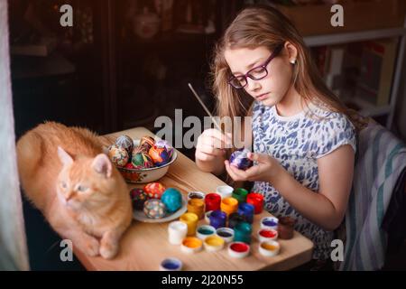 Teen girl malt Ostereier, ihre Ingwer-Katze liegt auf dem Tisch. Home Interieur, ostern Urlaub. Stockfoto