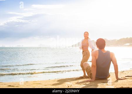 Ein junger Mann, der am Strand sitzt und eine junge Frau anschaut - Stockfoto Stockfoto