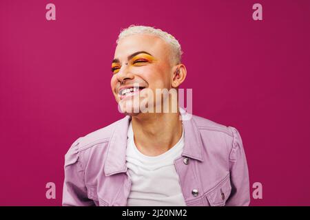 Queerer Mann, der fröhlich lächelt, während er in einem Studio steht. Unpassender junger Mann, der einen bunten Lidschatten vor einem violetten Hintergrund trägt. Frohe euch Stockfoto