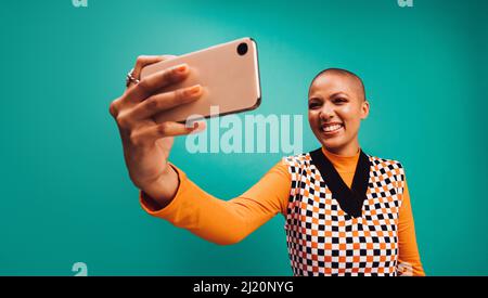 Lächelt für die Kamera. Glückliche junge Frau, die für ein Selbstporträt auf ihrer Smartphone-Kamera posiert. Junge Frau mit kurzen Haaren, die allein gegen eine tu steht Stockfoto