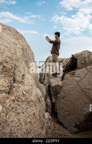 Junge Outdoor-Reisende, die Fotos von der Landschaft machen, während sie auf den Felsen am Strand stehen - Stock Photo Stockfoto