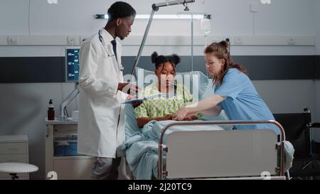 Allgemeinmediziner, die Gesundheitsberatung mit dem Patienten auf der Krankenhausstation durchführen. Facharzt diskutiert mit jungen Erwachsenen, während medizinische Assistentin hilft mit Oximeter, um Krankheit zu heilen Stockfoto