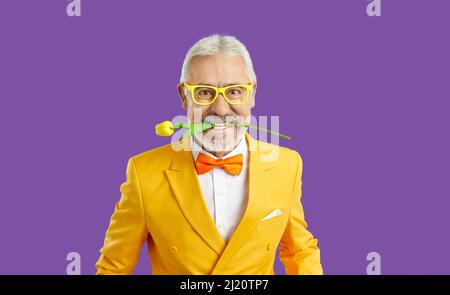 Fröhlicher, lustiger und exzentrischer älterer Mann mit frischer Tulpe in den Zähnen auf purpurem Hintergrund. Stockfoto