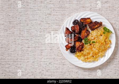 Geschmortes Sauerkraut mit Schweinefleisch und Pflaumen auf weißem Teller auf Leinentuch, horizontale Ansicht von oben, flaches Lay, Freiraum Stockfoto