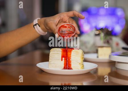 Weibliche Hand gießt Erdbeersoße über Crape Kuchen. Stockfoto