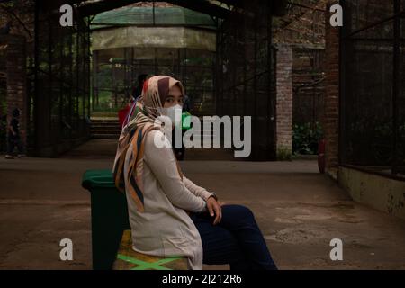 Schöne asiatische Mädchen, trägt eine Gesichtsmaske sitzen, in einem hellbraunen Kleid sitzen auf einem Stuhl im Gang Stockfoto