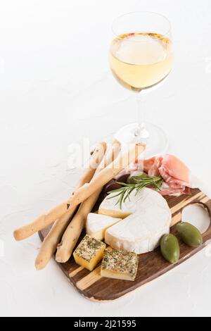 Holzkochplatte mit einer Auswahl an Käse und Prosciutto, serviert zusammen mit Oliven, Grissini und Wein auf einem Tisch aus weißem Stein. Italienische Traditionti Stockfoto