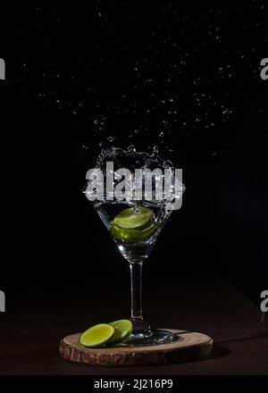 Eine vertikale Aufnahme eines Cocktails mit Limettenscheiben, in denen eine Limette ins Glas fällt und Flüssigkeit spritzt Stockfoto