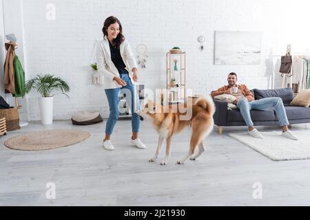 Lächelnde junge Frau spielt mit akita Inu Hund in der Nähe von glücklichen Freund mit einer Tasse Kaffee auf der Couch sitzen Stockfoto