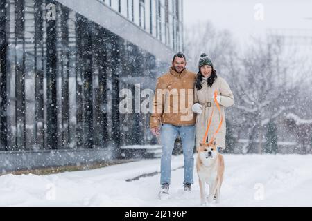 Lächelnde Frau, die Leine hält, während sie zusammen mit ihrem Freund und dem akita Inu-Hund unter fallendem Schnee läuft Stockfoto