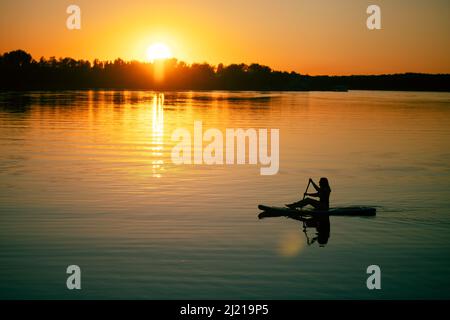 Frau Silhouette auf supboard mit einem Ruder in den Händen rudern auf erstaunlichen See mit ehrfurchtgebietenden Sonnenuntergang im Hintergrund bedeckt Wasseroberfläche mit gelb Stockfoto