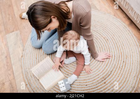 Ansicht von oben auf der Frau, die ein Buch liest, in der Nähe eines verschwommenen kleinen Sohnes auf dem Boden zu Hause Stockfoto