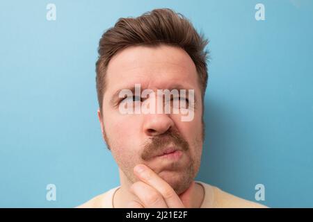 Mann mit lustigen Schnurrbart, der Zweifel hat und mit verwirrem Gesichtsausdruck auf isoliertem blauem Hintergrund Stockfoto