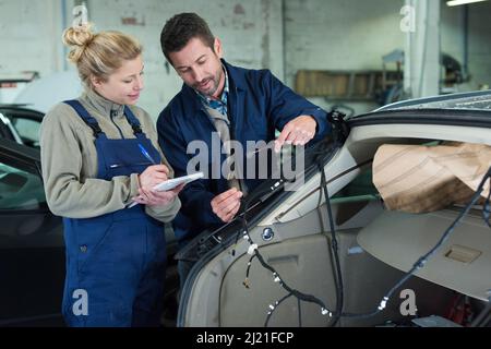 Junge Dame, die Notizen macht, während Mechaniker am Auto arbeitet Stockfoto