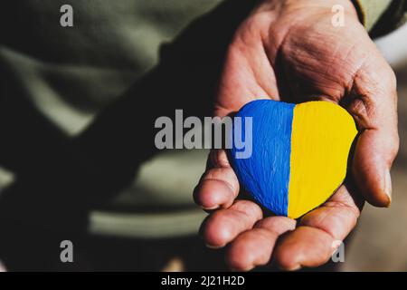 Ältere Mann oder Soldat Hände halten herzförmigen Stein mit Ukraine Nationalflagge Farben gemalt. Betet für die Ukraine, beendet den Krieg, rettet die Ukraine und ihre Kinder. Keine Invasion, Besetzung und Terrorismus Stockfoto