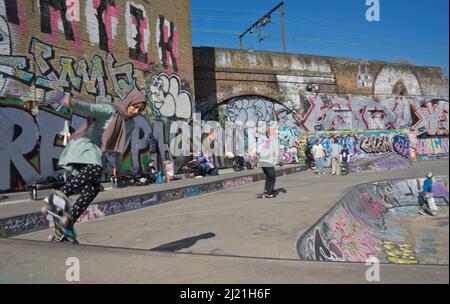 Junge muslimische Frau, die mit dem Zug und dem überirdischen Bahnhof in Tower Hamlets, London, England, Großbritannien, unter Eisenbahnbögen Schlittschuhlaufen kann Stockfoto