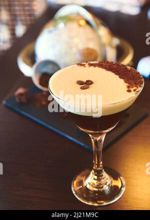 Ein Espresso martini mit Kaffeebohnen und Schokoladenstaub, der vor Kaffeebohnen und einem Globus zu sehen ist Stockfoto